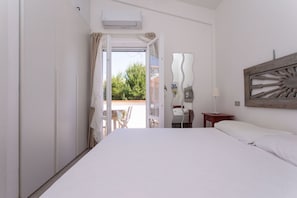 A bedroom with a bed, a desk, a chair, and an open door leading to an outdoor area.