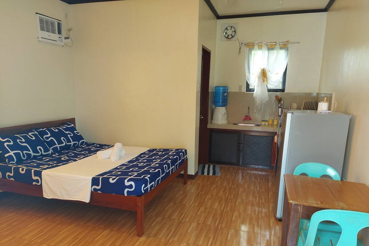Una habitación compacta con cama, kitchenette pequeño, mesa y silla.