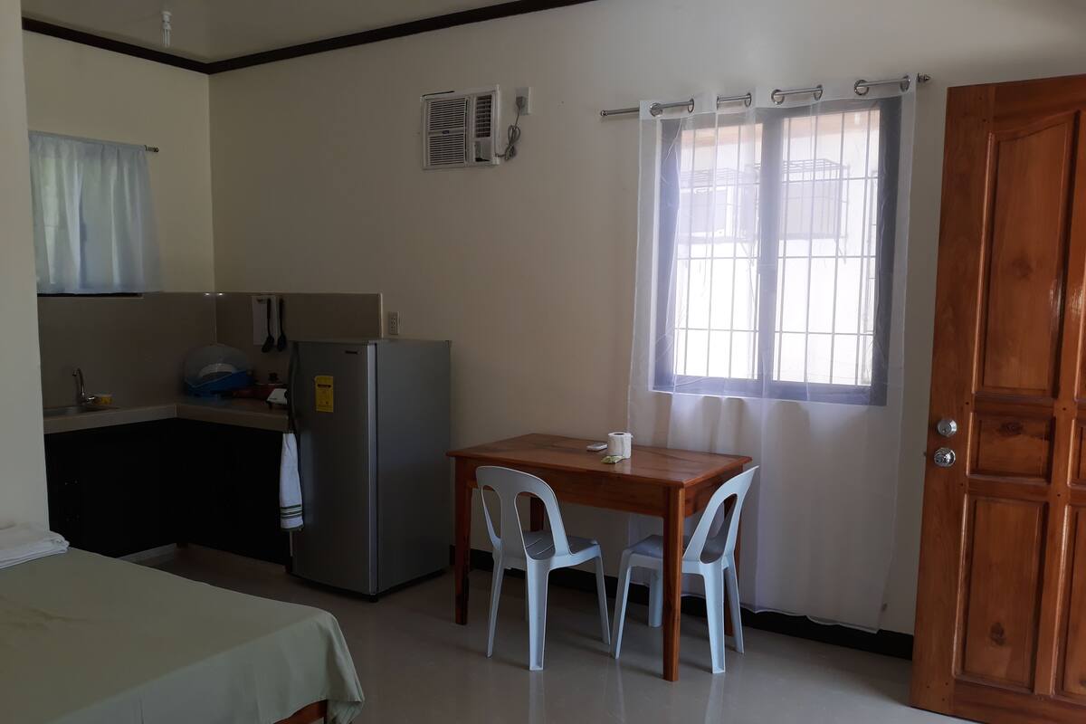 Una habitación pequeña y limpia con cama, kitchenette, una mesa con sillas y ventana con cortinas.