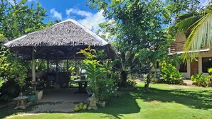 A thatched-roof pavilion in a lush garden with a palm tree and a building in the background. - Gardenview Homestay Siargao (General Luna)