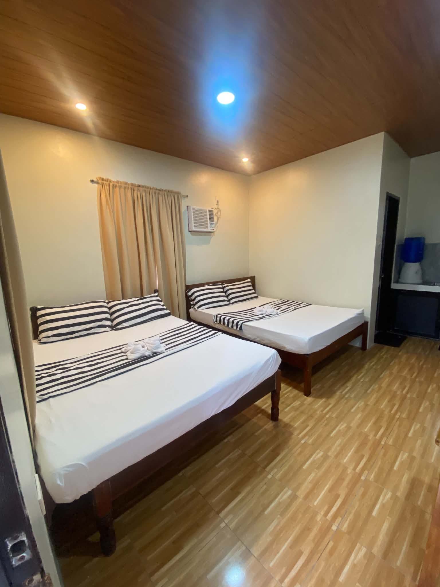 A hotel room with two beds, wooden flooring, a ceiling light, and a window with curtains.