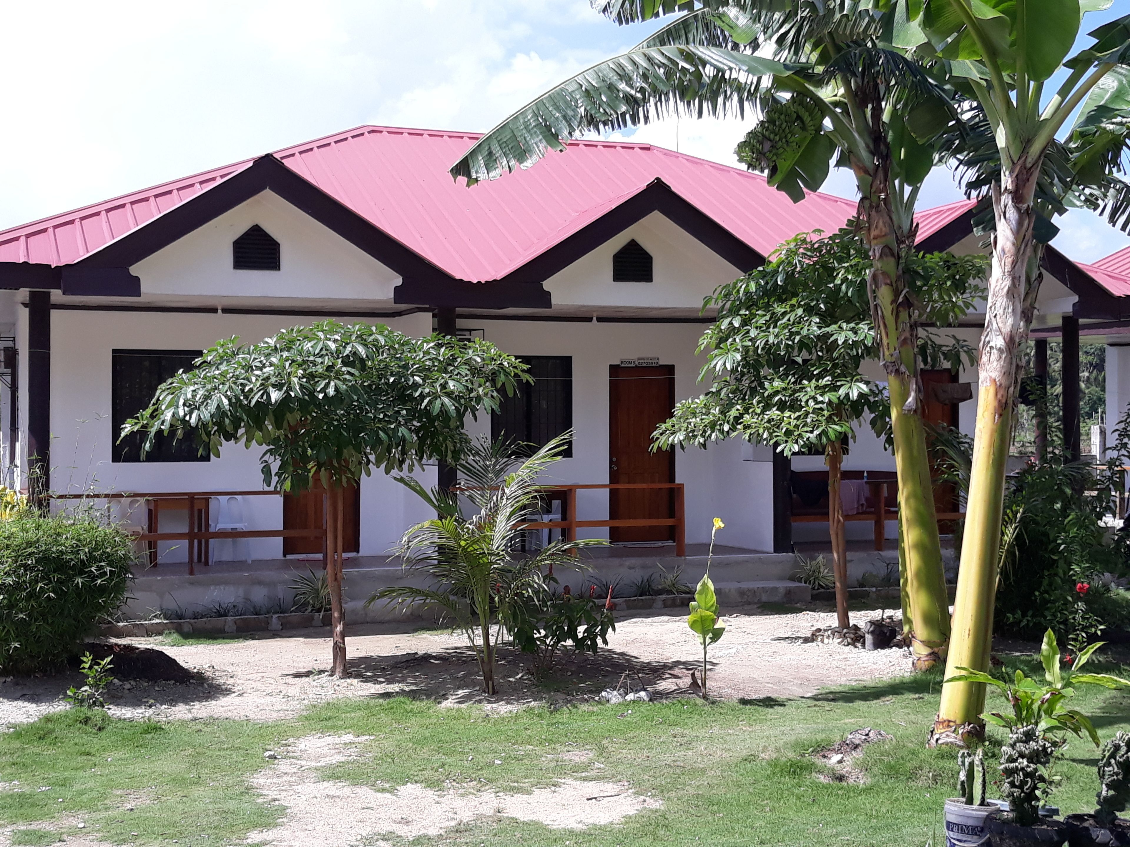 A building with a red roof and white walls, surrounded by a garden with various plants and a bench.