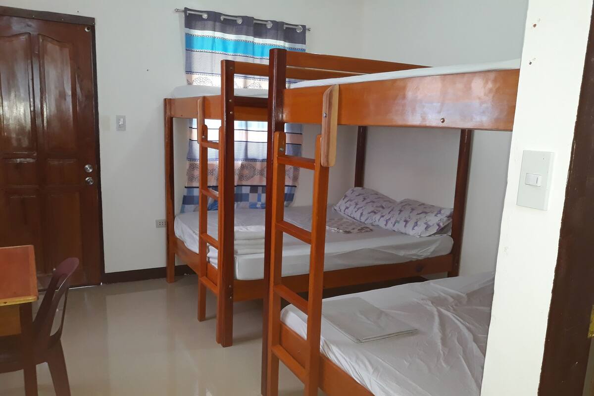 Una habitación con dos literas, una puerta de madera, una silla y un ventanal con cortinas.