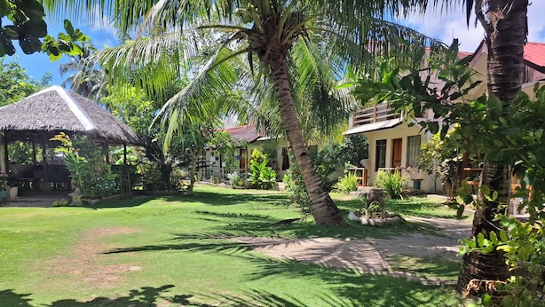 A tropical garden with palm trees, a thatched-roof structure, and a pathway leading to a building with a red roof. - Gardenview Homestay Siargao (General Luna)