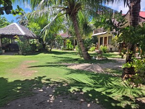 A tropical garden with palm trees, a thatched-roof structure, and a pathway leading to a building with a red roof. - Gardenview Homestay Siargao (General Luna)