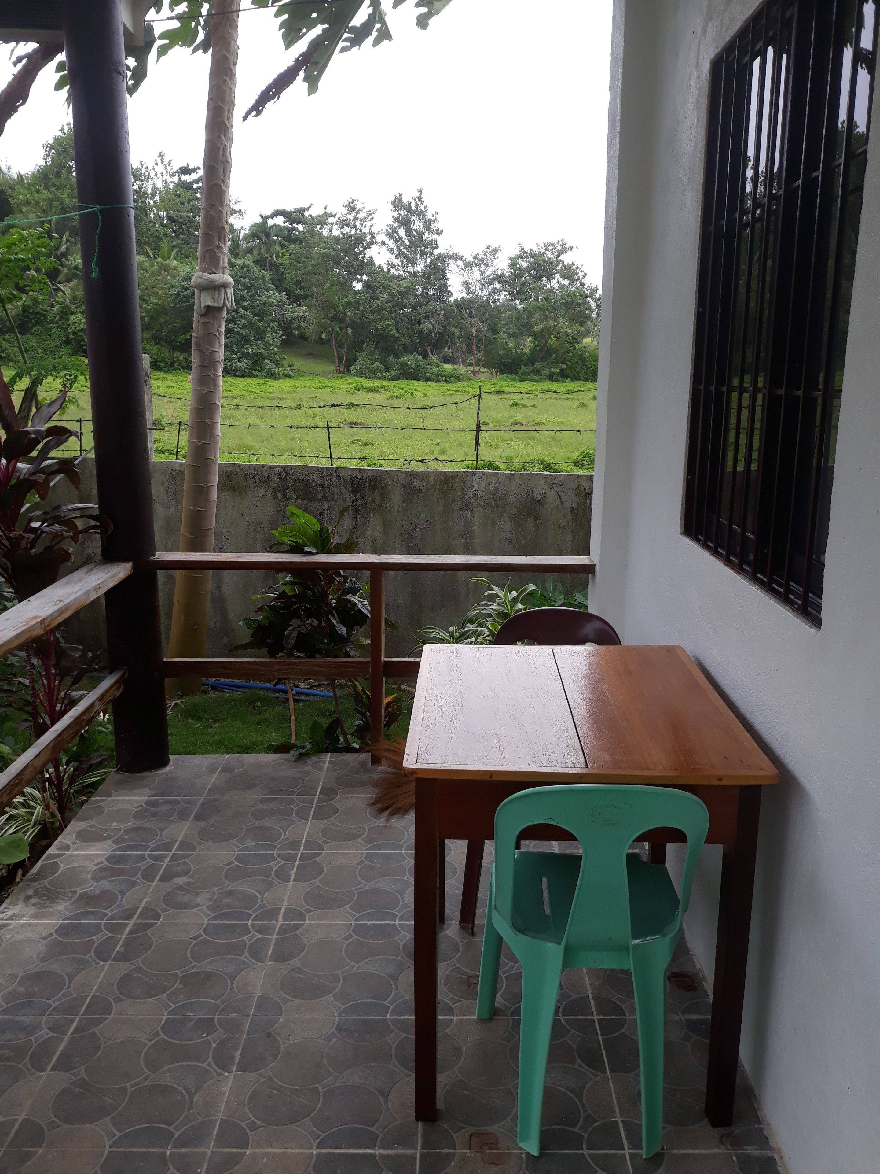 Un patio con una mesa de madera y una silla de plástico verde, un muro de hormigón y vistas a un campo verde y árboles.