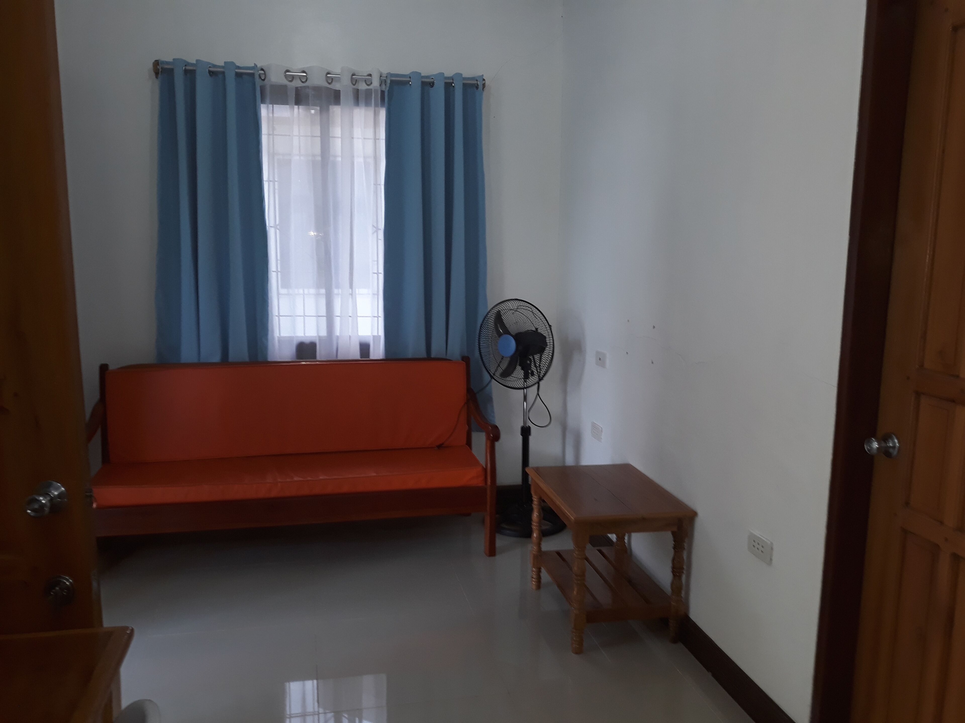 A room with a wooden sofa, a small wooden table, a standing fan, and a window with blue curtains.