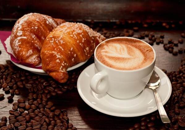 Une tasse de café ornée d’un motif en latte, posée sur une soucoupe, un croissant saupoudré de sucre sur une assiette, et quelques grains de café éparpillés.