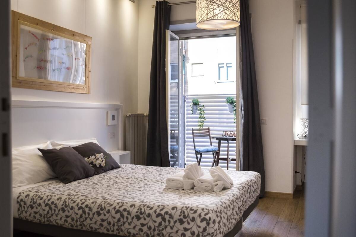 A neatly made bed with a patterned bedspread, a framed map on the wall, and a view of the outside balcony through the open door.