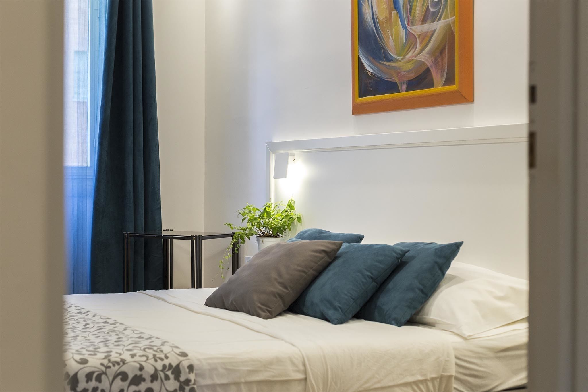 A neatly made bed with white and blue bedding, a bedside plant, and a painting on the wall.