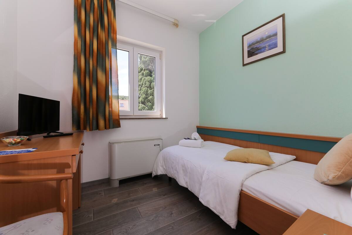 A bedroom with a bed, a desk, a chair, a window with curtains, a radiator, and a framed picture on the wall.