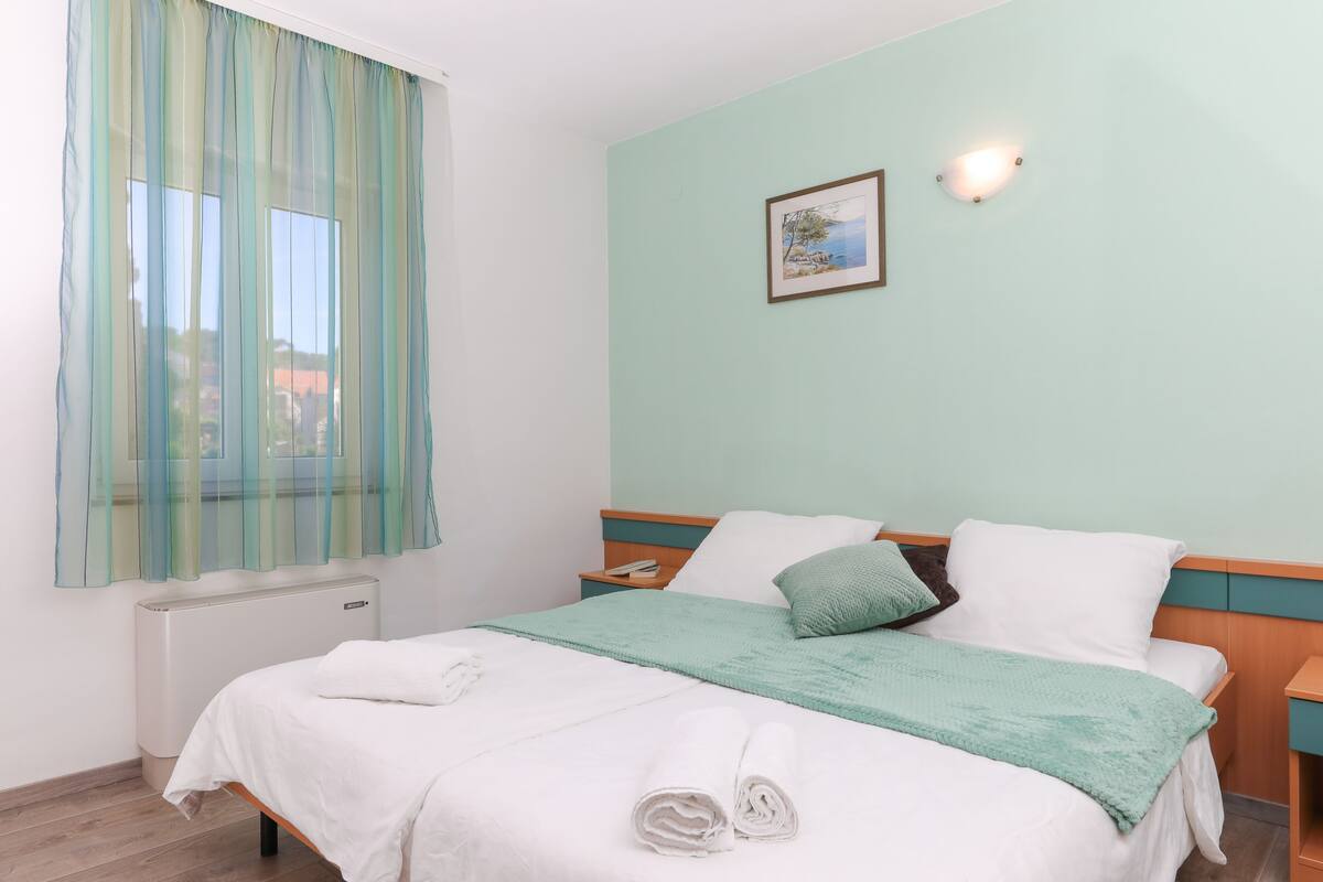 A neatly made bed with white linens and a green throw blanket, a bedside table with a lamp, and a window with sheer curtains.