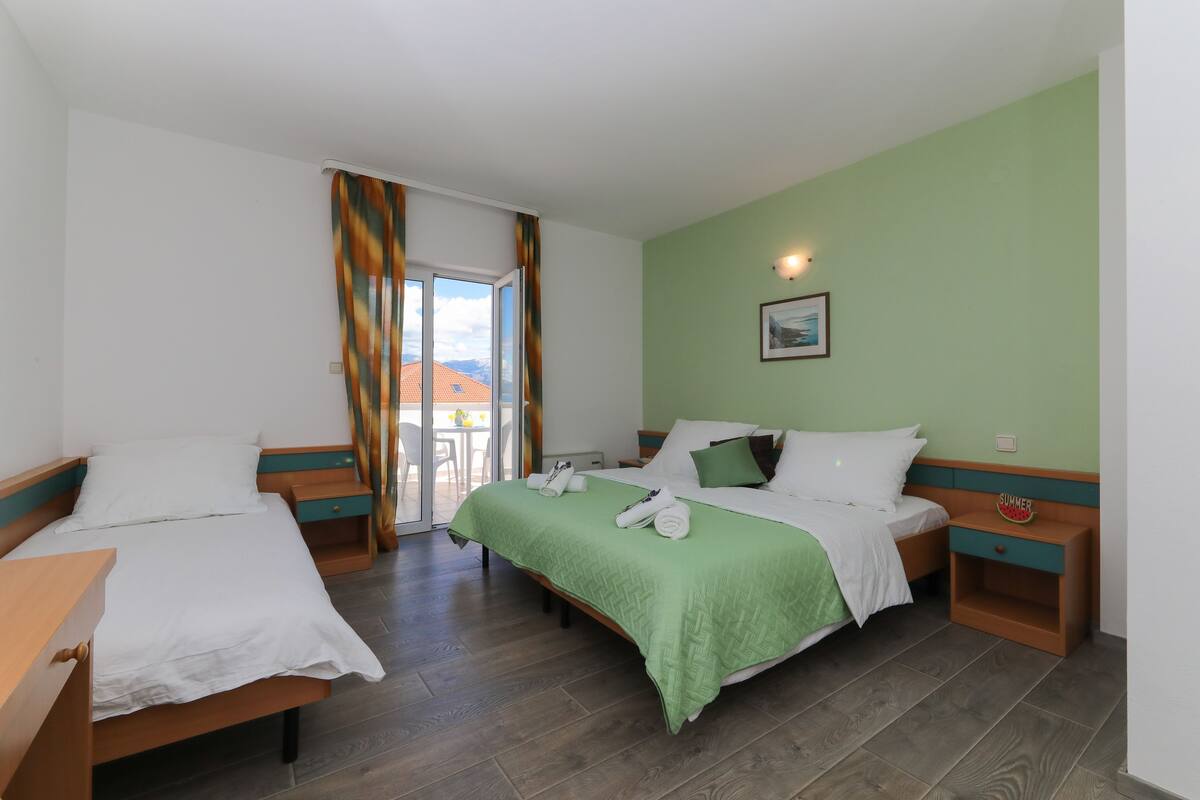 A hotel room with two beds, a green wall, a picture, and a view of the outdoors.