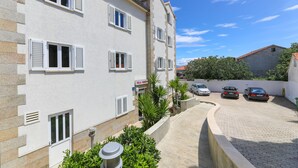 Un immeuble à plusieurs étages aux murs blancs et aux volets fermés, avec une allée pavée et des voitures garées.