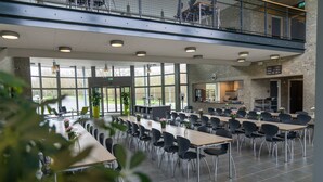 A spacious dining area with long tables and chairs, large windows, and a kitchen area visible through an open door. - ICV SleepWell (Vra)