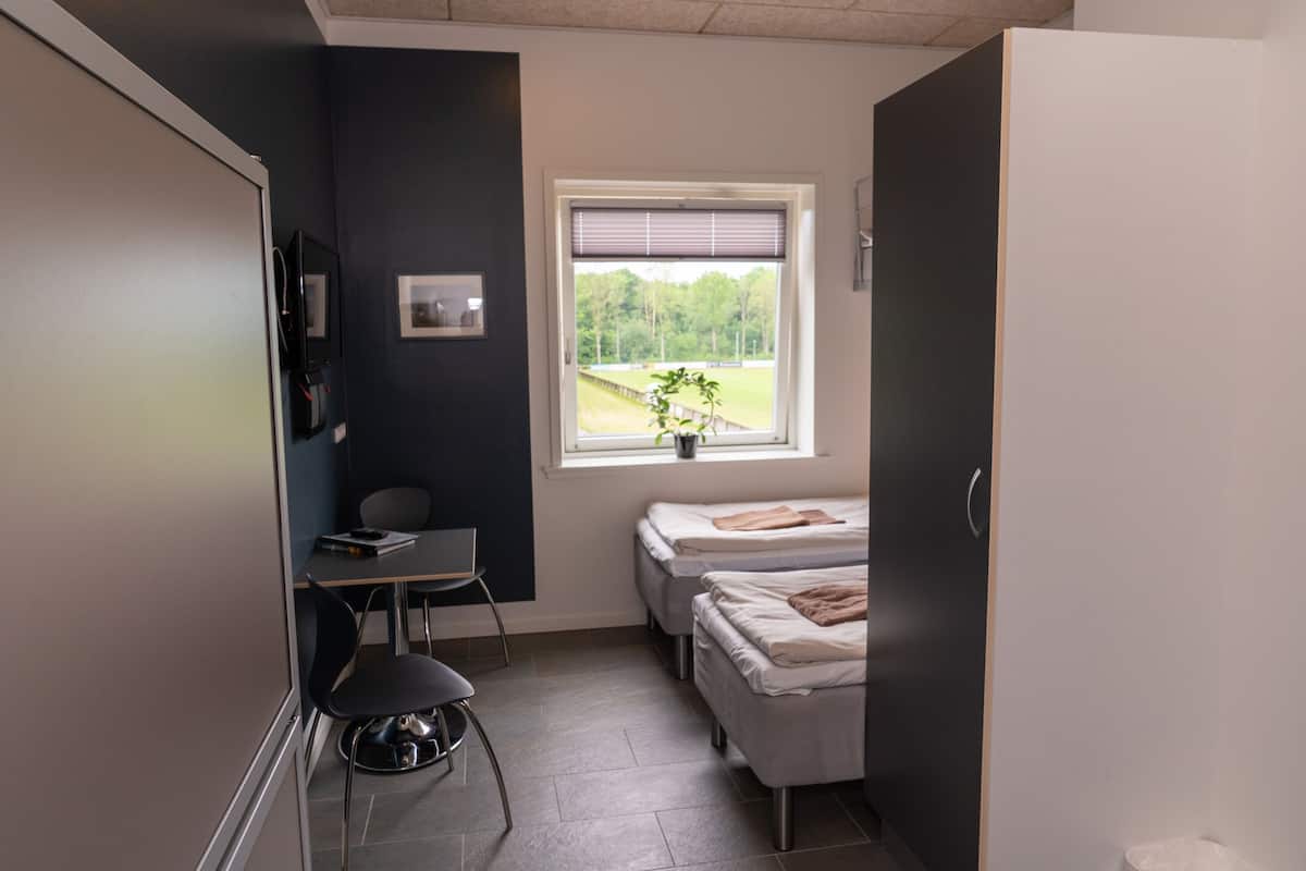 A small, modern room with a desk, two single beds, a window with a view of a field, and a door.