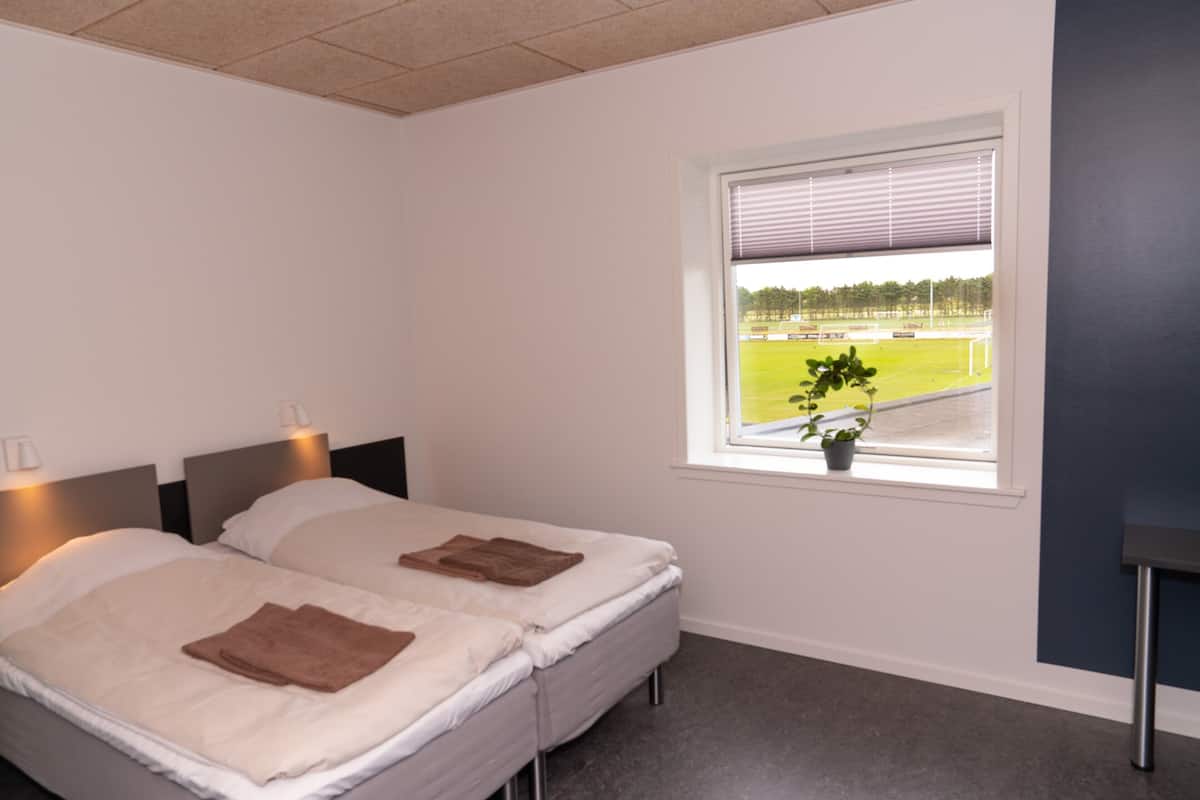 A bedroom with two beds, a window with a view of a sports field, and a small potted plant on the windowsill.