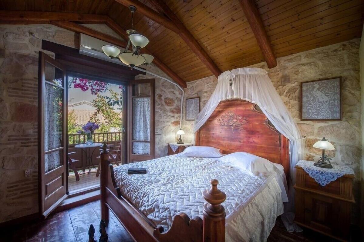 A bedroom with a wooden bed, a stone wall, a balcony with a table and chairs, and a view of the outside.