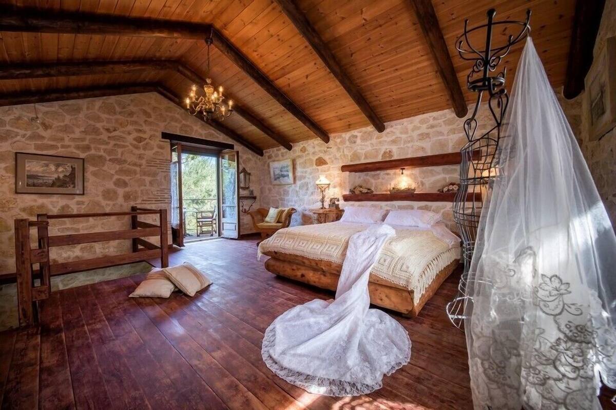 A rustic bedroom with a stone wall, wooden beams, a bed with a canopy, and a wooden chair.