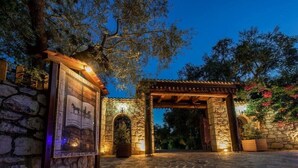 A stone entrance with a wooden archway, a framed sign, and a pergola with lights. - Ktima Kourou (Zakynthos)
