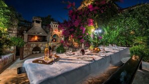 An outdoor dining area with a stone fireplace, a long table set for a meal, and lush greenery. - Ktima Kourou (Zakynthos)