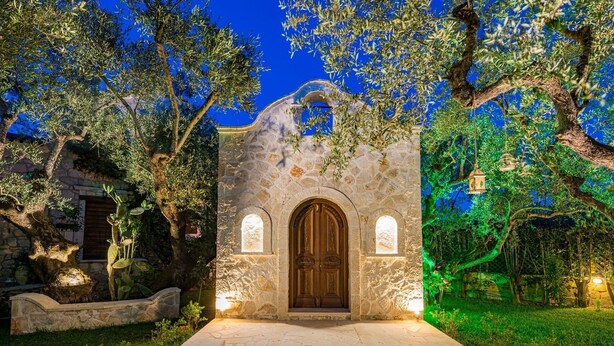 A stone building with a wooden door and arched windows, surrounded by greenery and stone walls. - Ktima Kourou (Zakynthos)