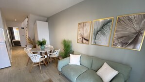 Un salon moderne avec un canapé gris, des coussins décoratifs blancs et une table de salle à manger ronde avec des chaises blanches. Trois œuvres d’art encadrées sont accrochées au mur au-dessus du canapé.