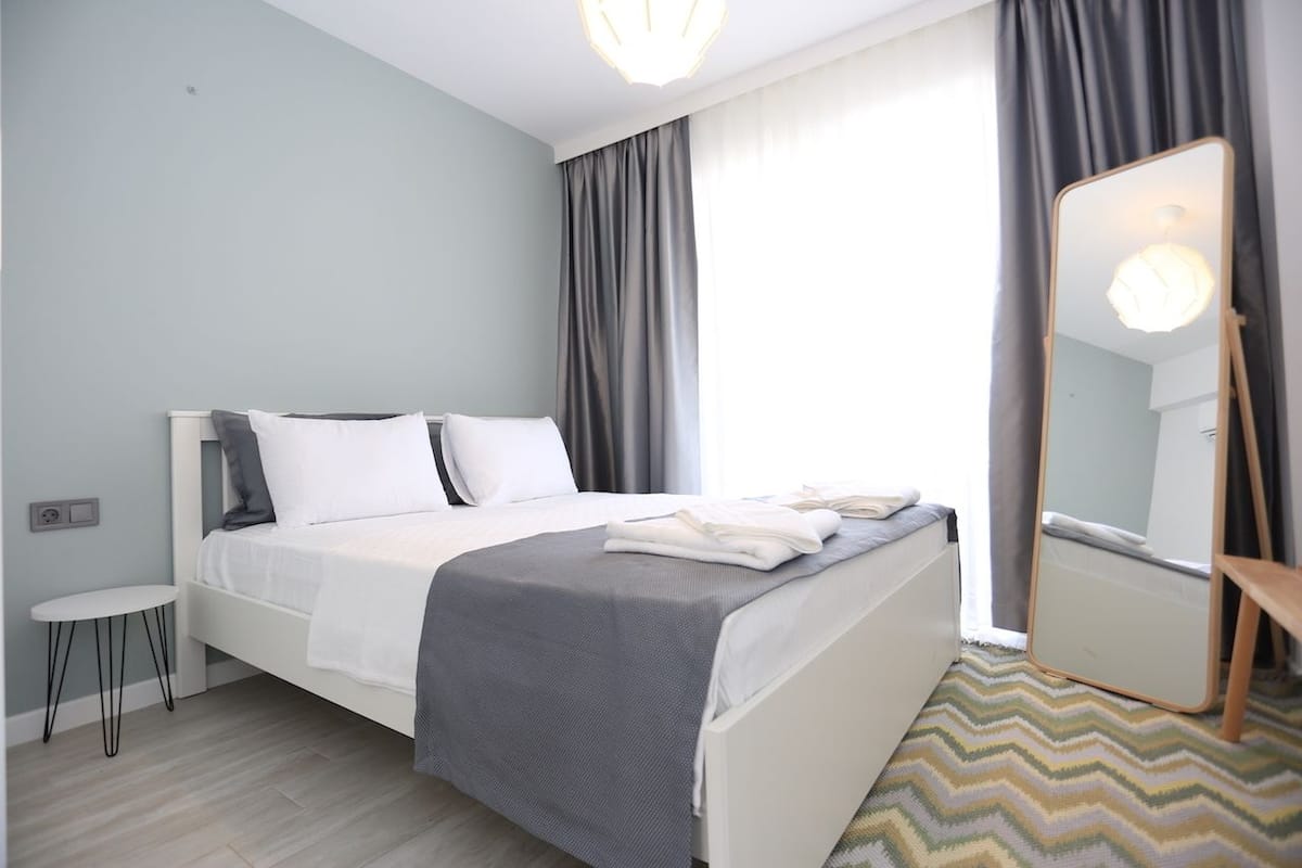 A modern bedroom with a bed, a small table, a mirror, and a view of the outside through a window.