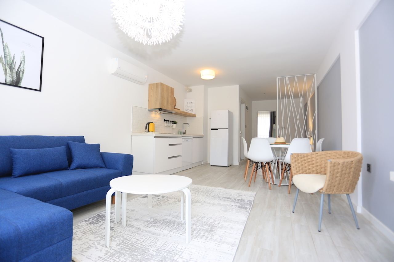 Una sala de estar moderna con un sofá azul, una mesa de centro blanca y un comedor con sillas blancas y una silla de mimbre.