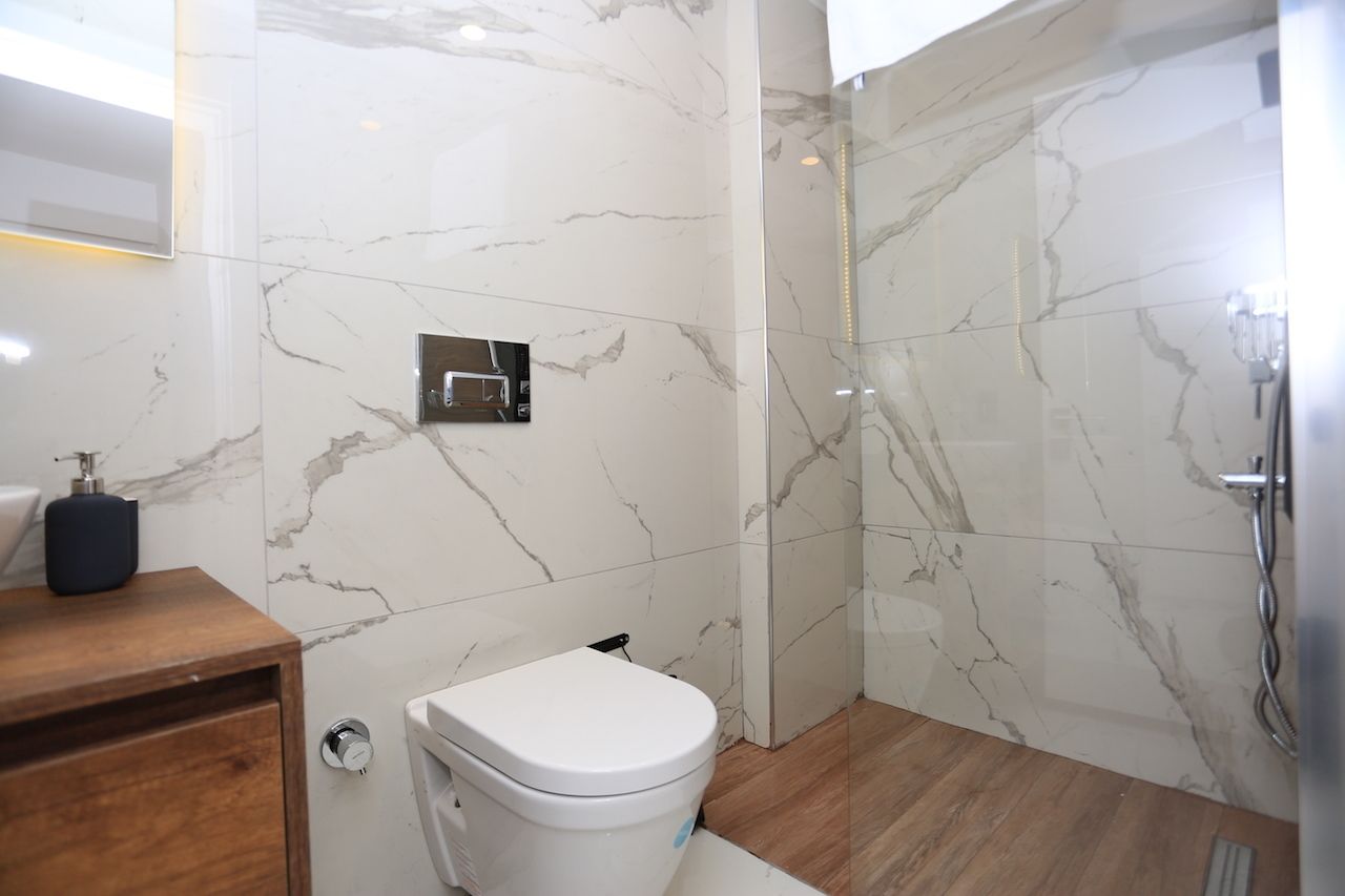 Et moderne badeværelse med et marmorgulv, et hvidt toilet, et træskab og en glasbrusekabine.
