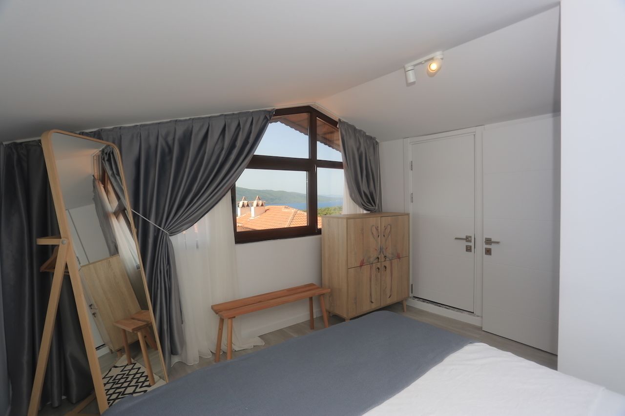 A bedroom with a bed, a wooden wardrobe, a bench, a mirror, and a window with curtains.