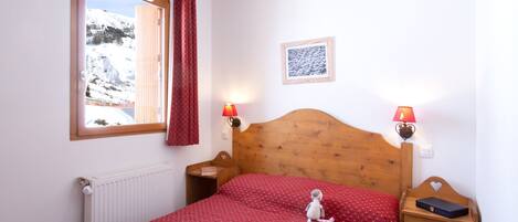 A bedroom with a wooden headboard, a red patterned bedspread, two bedside lamps, a framed picture on the wall, and a window with a view of a snowy landscape.