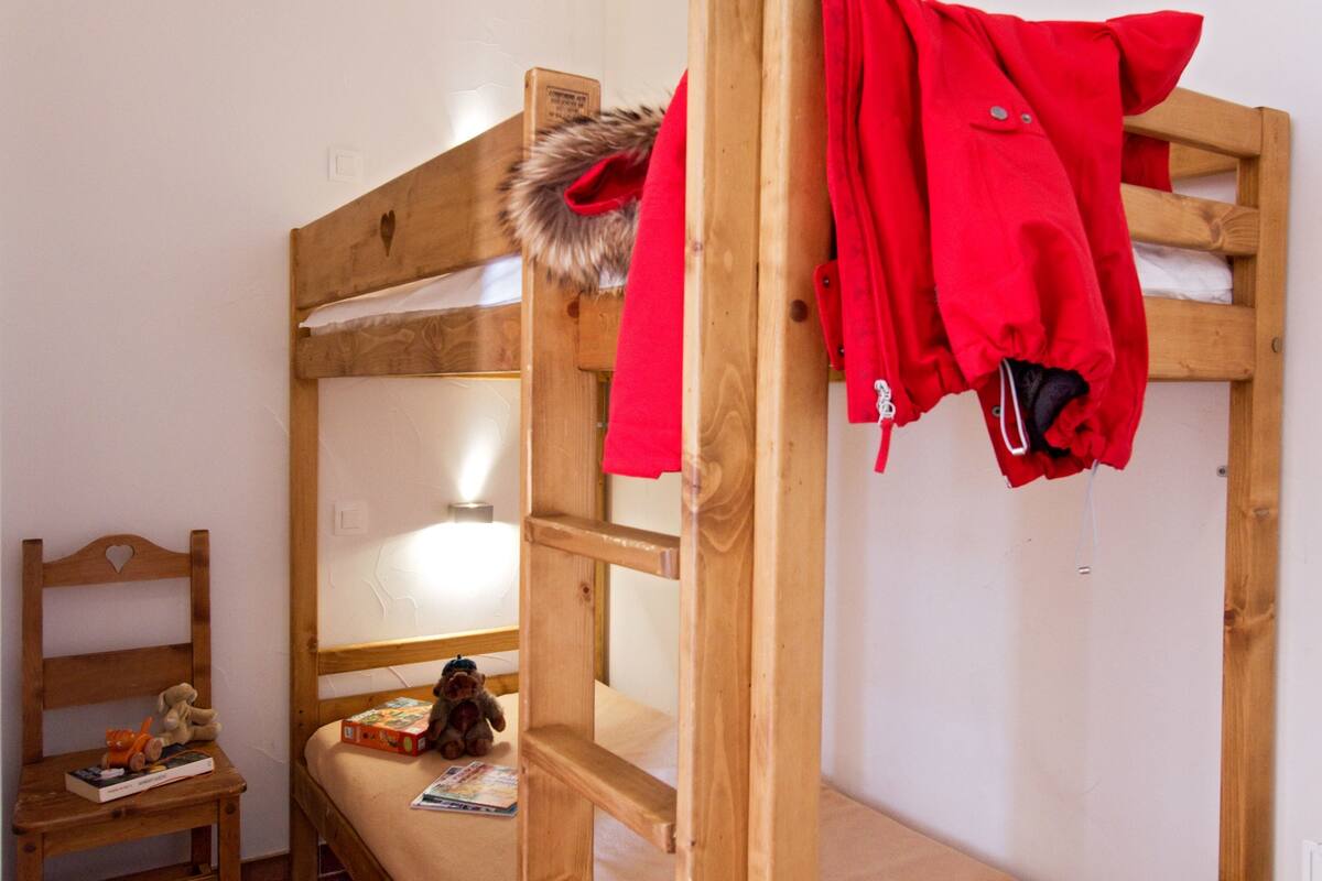 Una cama litera con cabecera de madera, una pequeña silla de madera y un estante con libros y un peluche.