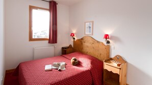 A bedroom with a wooden headboard, a red bedspread, a small round rug, a book, a lamp, and a chessboard on a wooden nightstand.