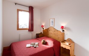 A bedroom with a wooden headboard, a red bedspread, a small round rug, a book, a lamp, and a chessboard on a wooden nightstand.