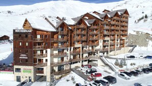 A multi-story wooden hotel with balconies, a ski lift in the background, and a parking area.