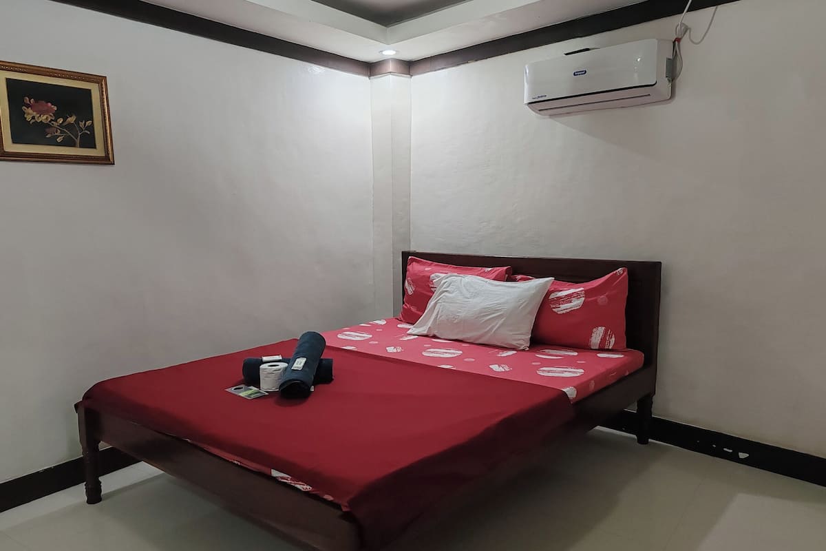 Un lit bien fait, avec une couverture rouge et des oreillers, un tableau encadré au mur et un appareil de climatisation.