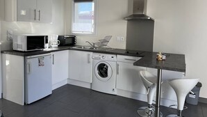 Cocina compacta con armarios blancos, microondas, cafetera, lavadora y un lavabo.