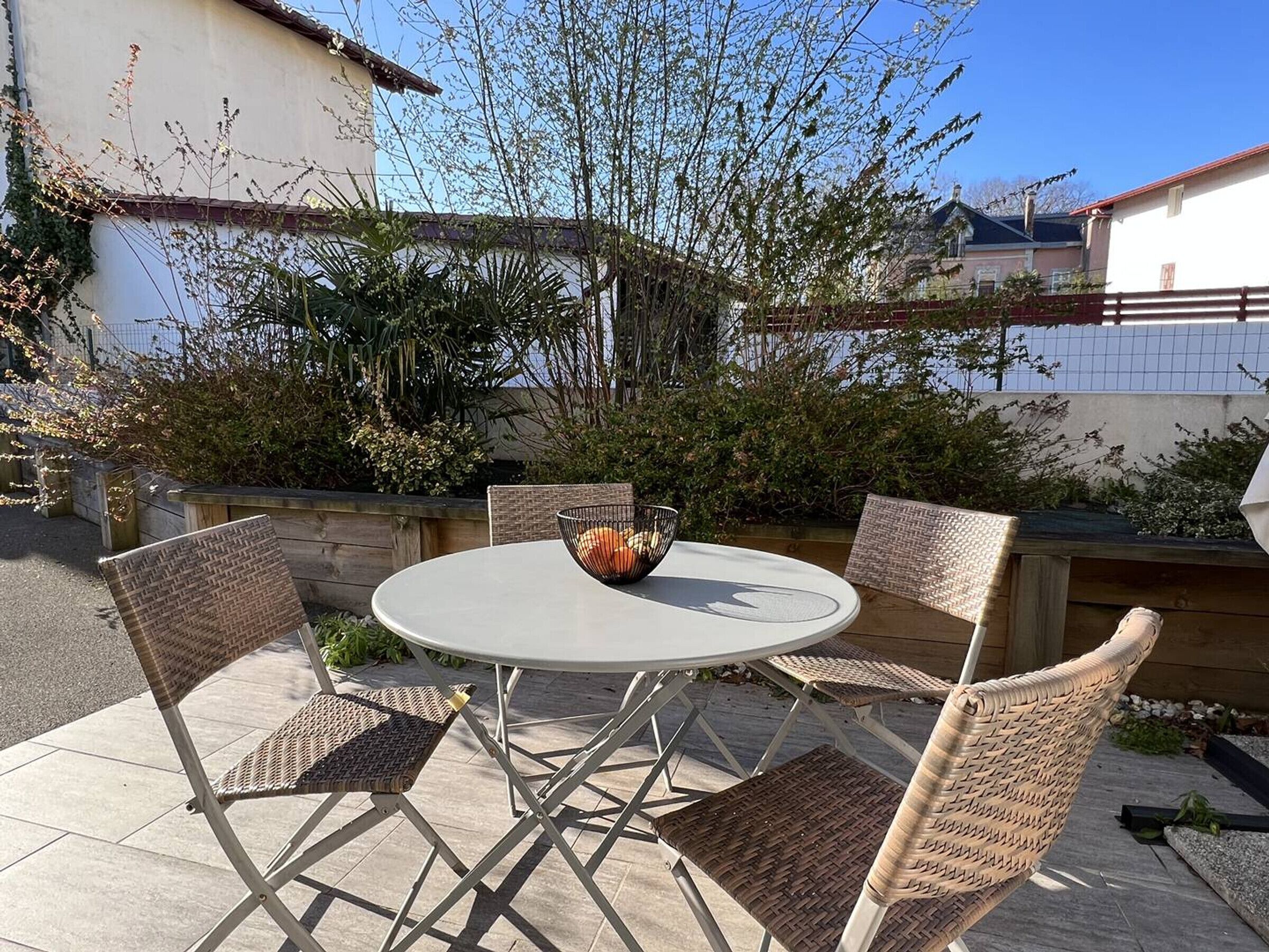 An outdoor patio with a round table and four wicker chairs, surrounded by greenery and a white building.