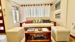A living room with a sofa set, a coffee table with books, a window with curtains, and a wall with framed pictures.