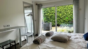 A bedroom with a large bed, a small table, a chair, and a view of a garden through a sliding door. - Ecocoon Cottage – Sleeps 4 – 2 Bathrooms – Heated Indoor Pool (Arques)