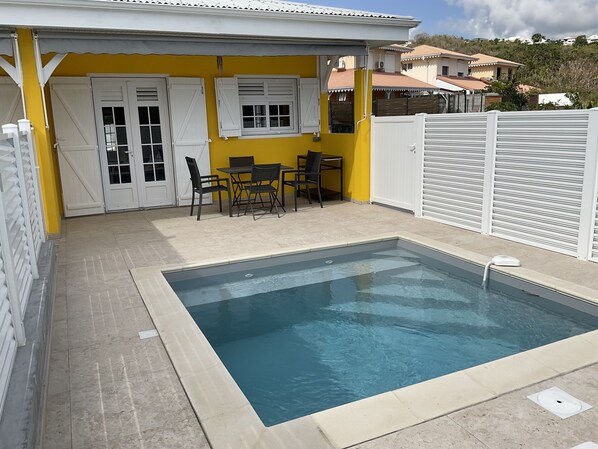 Ein Schwimmbecken mit Fliesenränderung, einem Tisch mit Stühlen und einer gelben Wand mit weißen Fensterläden.