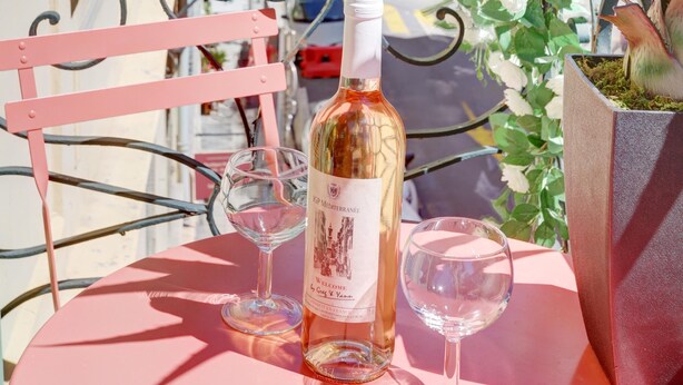 Een tafel met een fles wijn en twee glazen, een bloempot en een roze stoel op een balkon met uitzicht op een straat vol kleurrijke gebouwen.