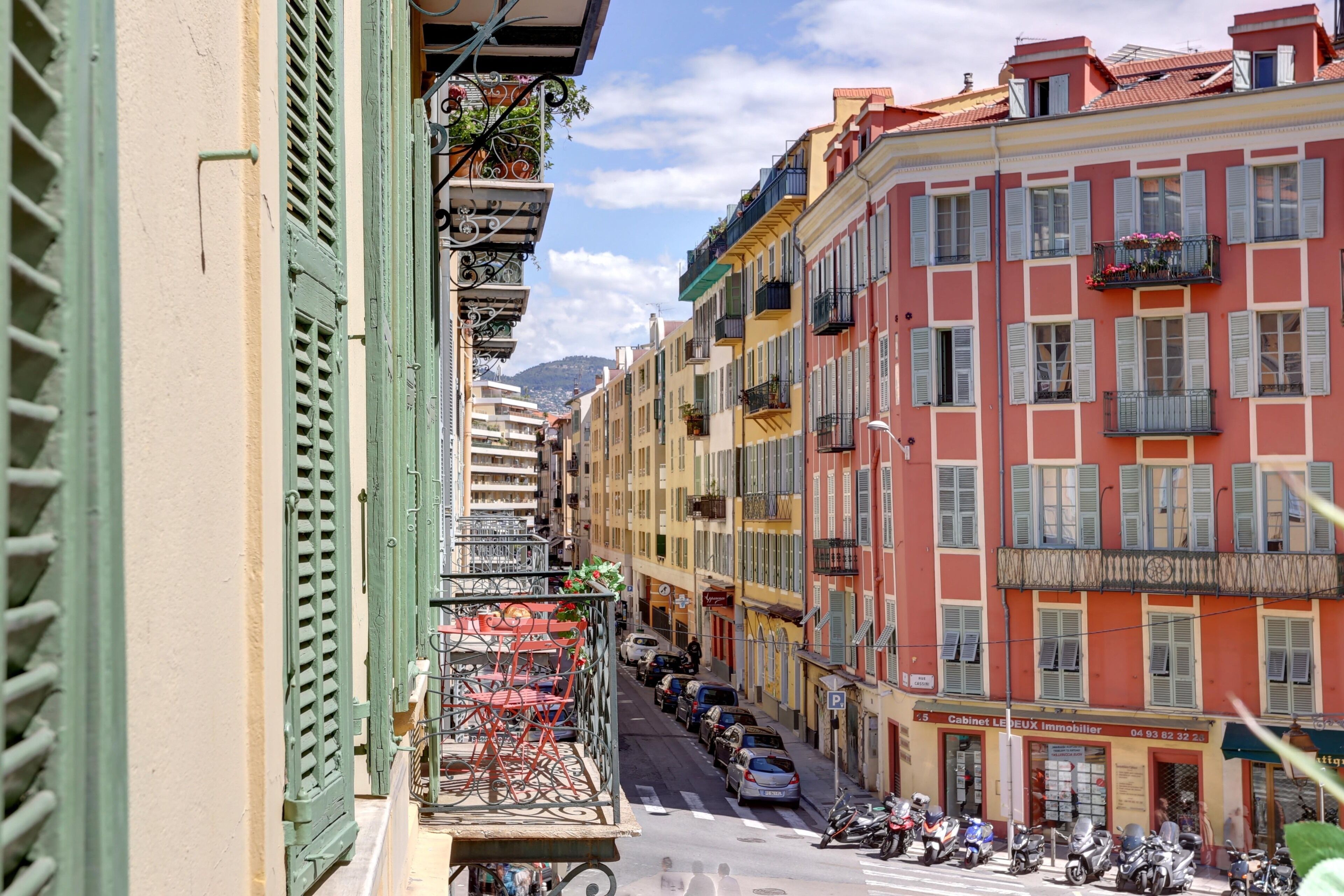 Une rue étroite bordée de bâtiments aux couleurs vives, de balcons ornés de plantes et de voitures garées.