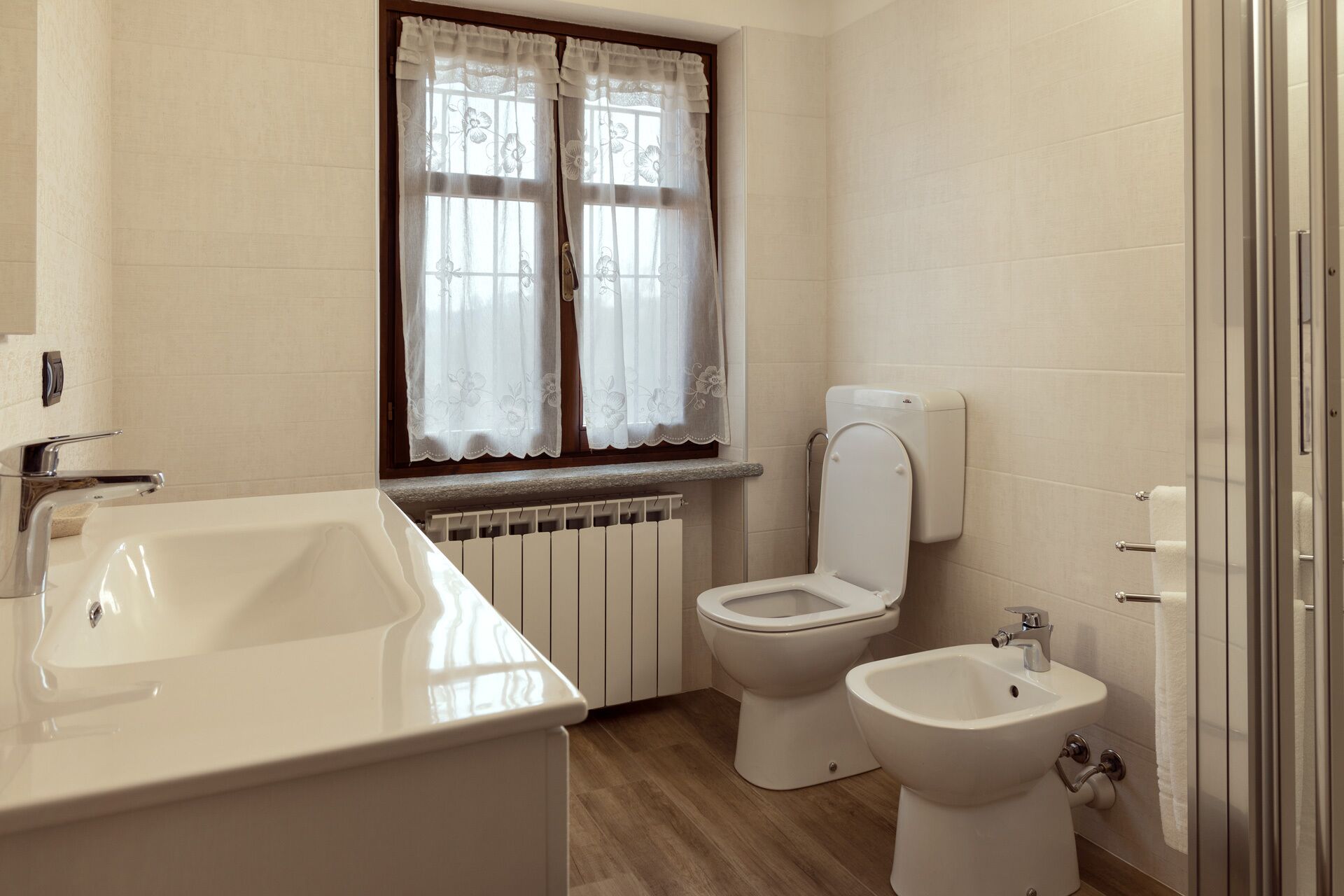 A bathroom with a white sink, toilet, and bidet, a window with floral curtains, and a radiator.