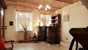 Una habitación con techo de ladrillo, dos ventanas con cortinas estampadas, un banco de madera, una mesita con una lámpara y una estantería.