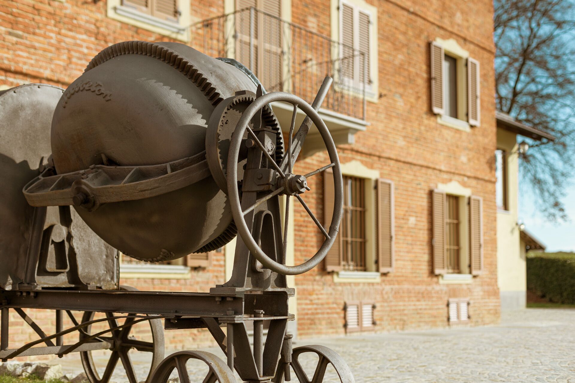 A cement mixer with a large wheel and a metal frame, positioned in front of a brick building with windows and shutters.