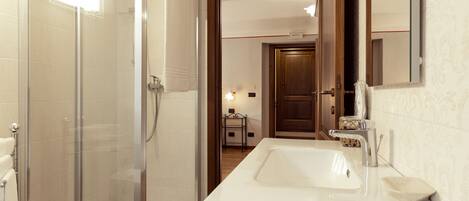 Un baño moderno con ducha de vidrio, un lavamanos blanco con jabonera y una puerta de madera que da a otra habitación.