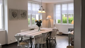 Een eetruimte met een lange tafel, witte stoelen en een bloevas. Er hangen ingelijste foto's aan de muur en staat een lamp op tafel.