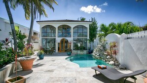 A two-story house with a swimming pool, surrounded by tropical plants and a stone patio.